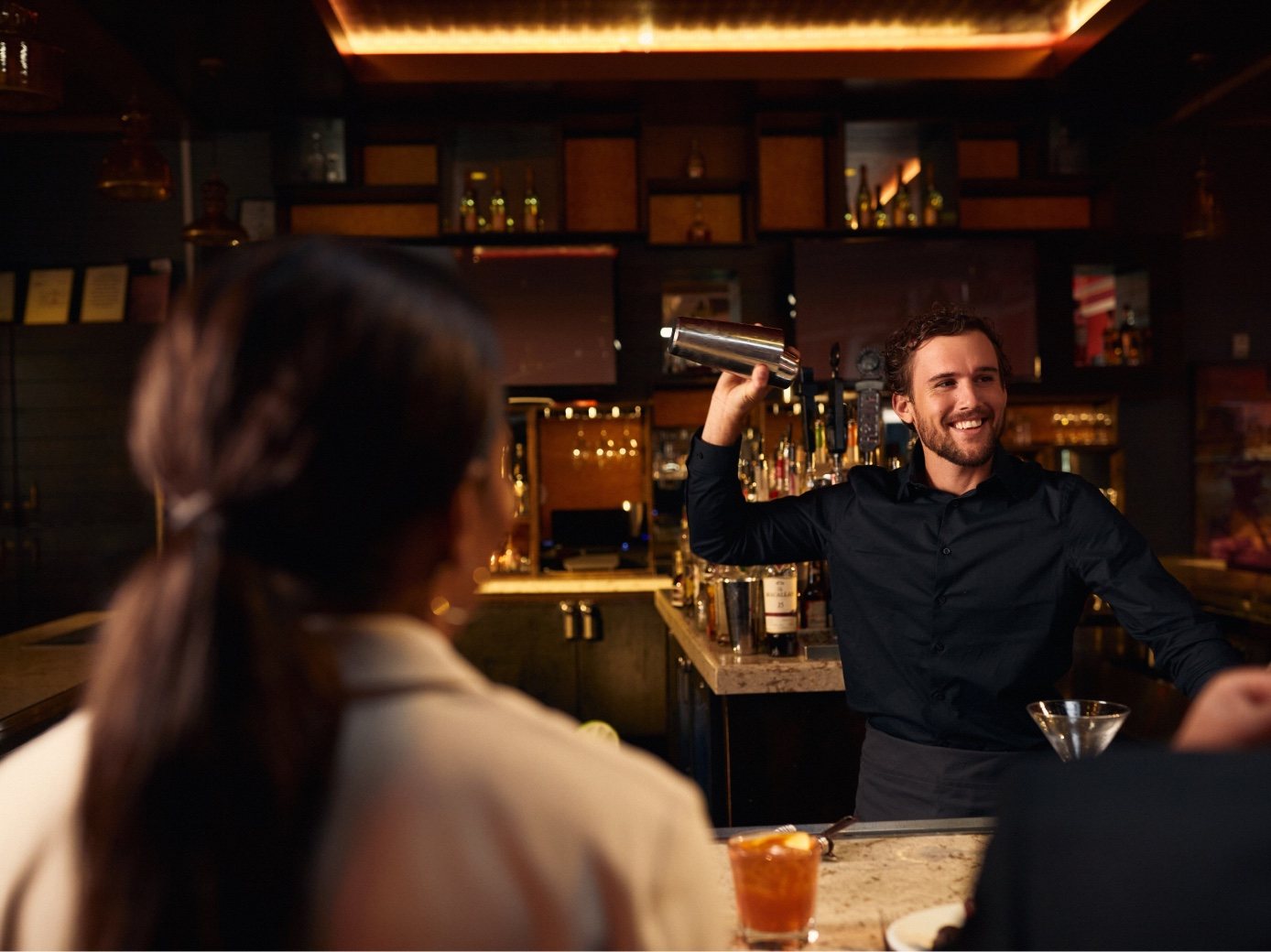 A bartender shakes a cocktail and grins at a guest across the bar