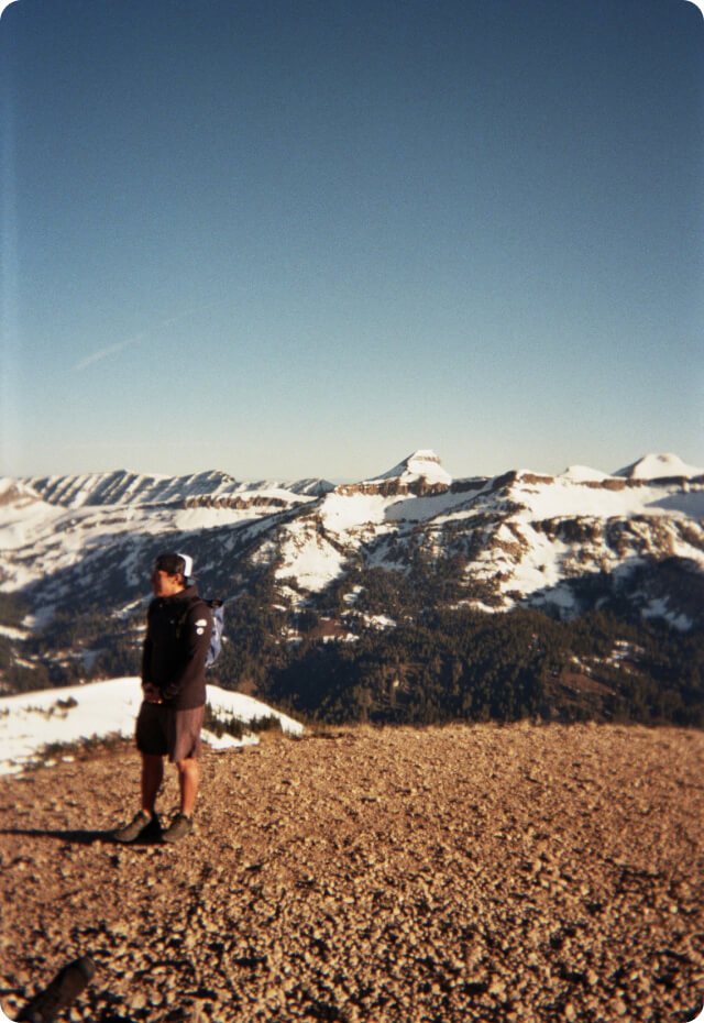 A crew member standing on a rocky summit above snow-covered peaks