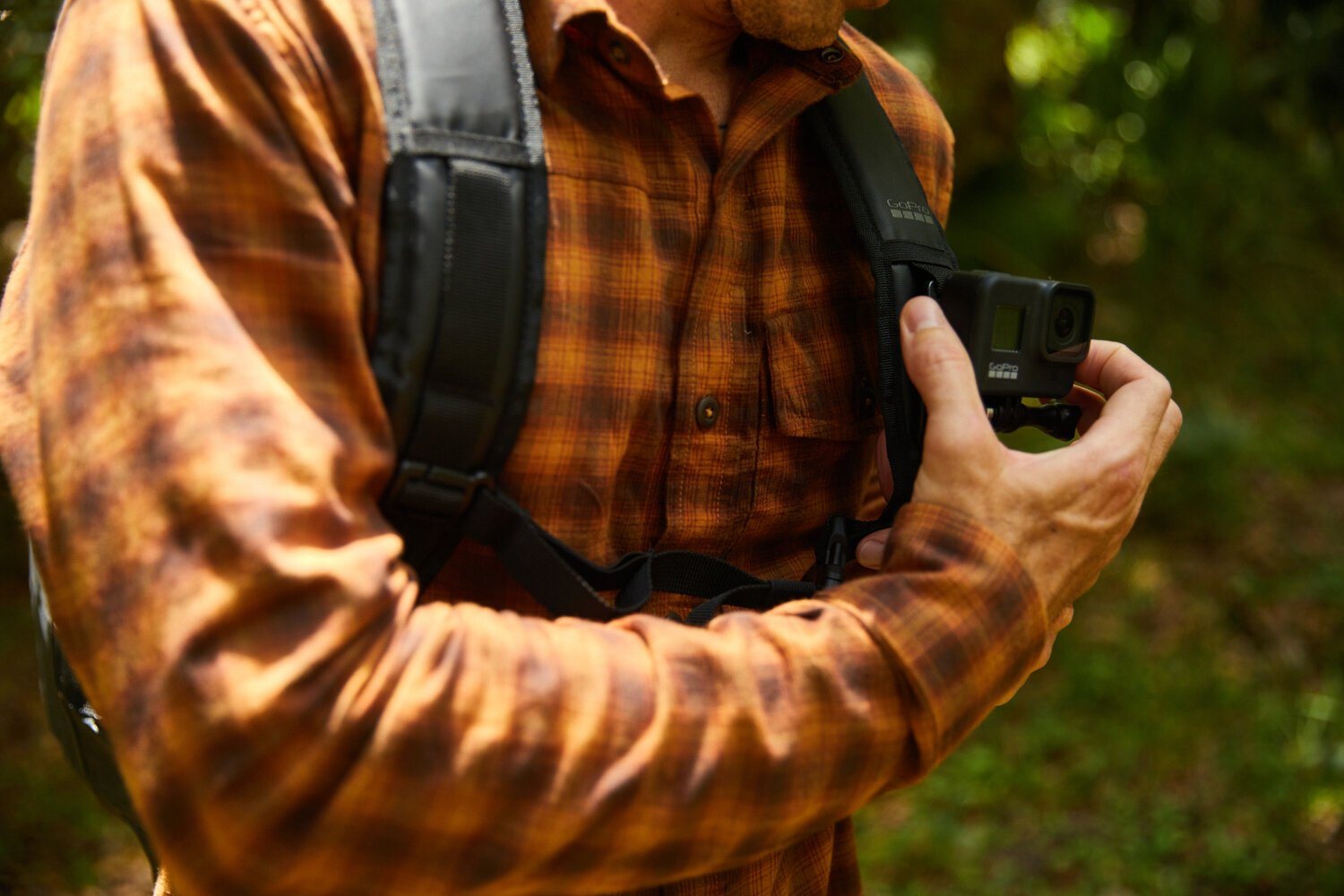 A hiker in a plaid shirt clips a GoPro camera to his backpack strap