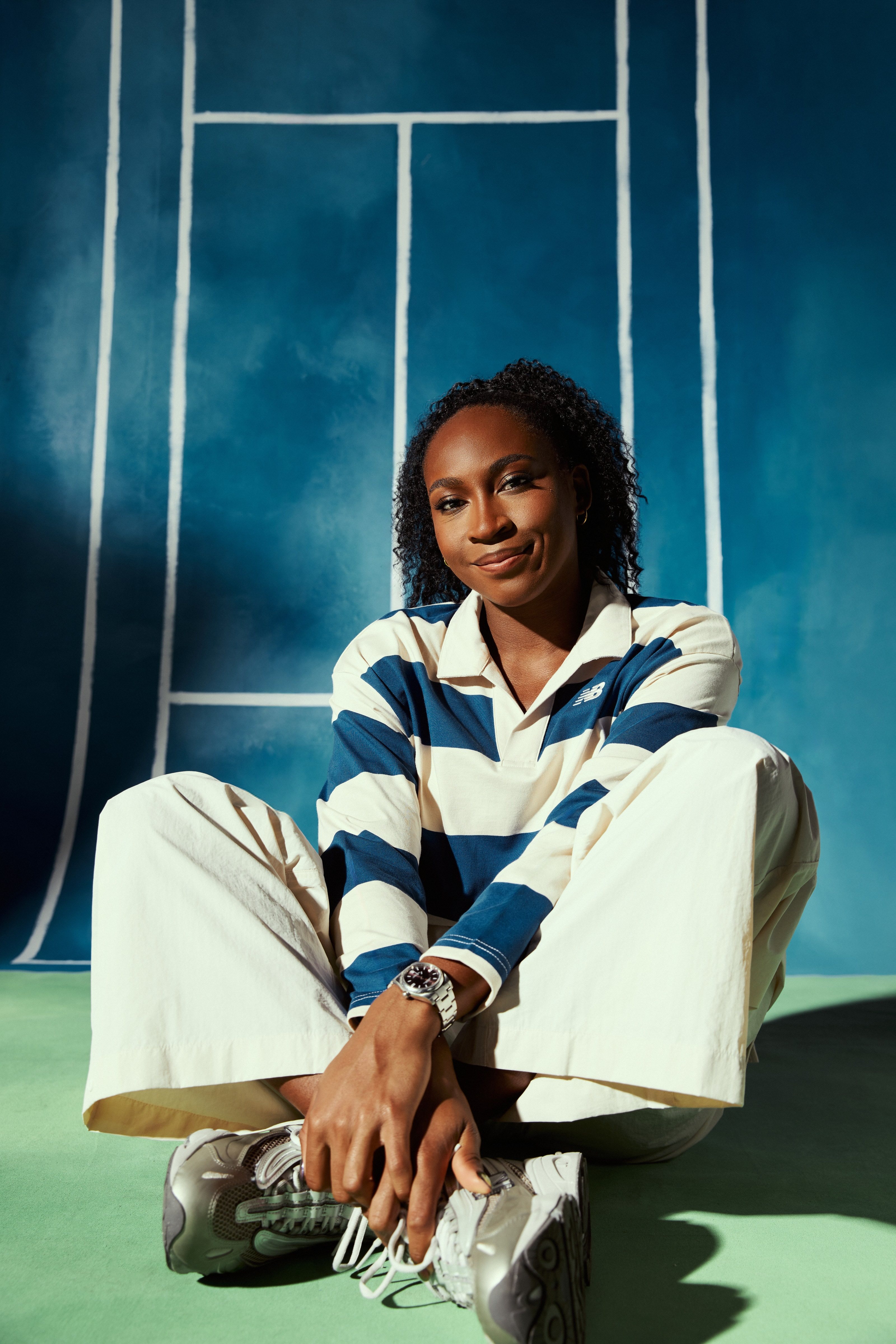 Coco Gauff smiles from the floor in a striped rugby shirt against a painted blue court