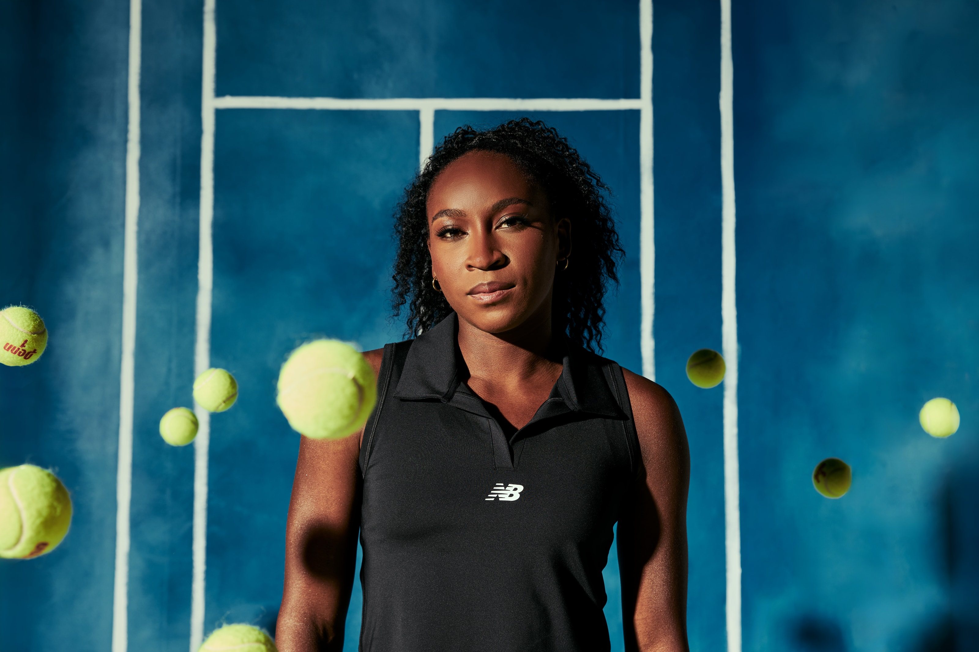 Coco Gauff stands still in a black polo as tennis balls fly past against a painted blue court