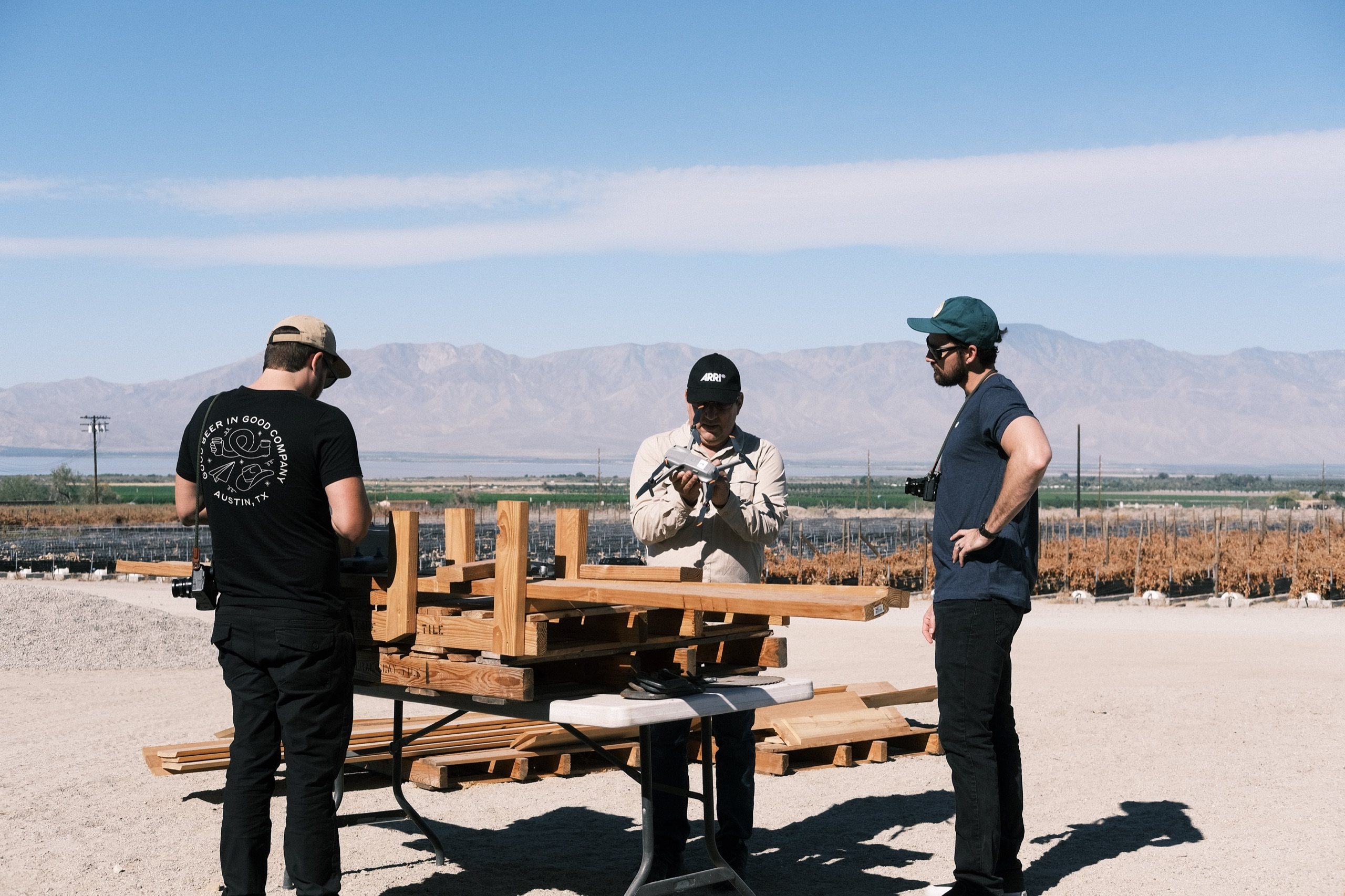 Two Spry crew members with cameras talk with a man at a table on a farm, mountains behind