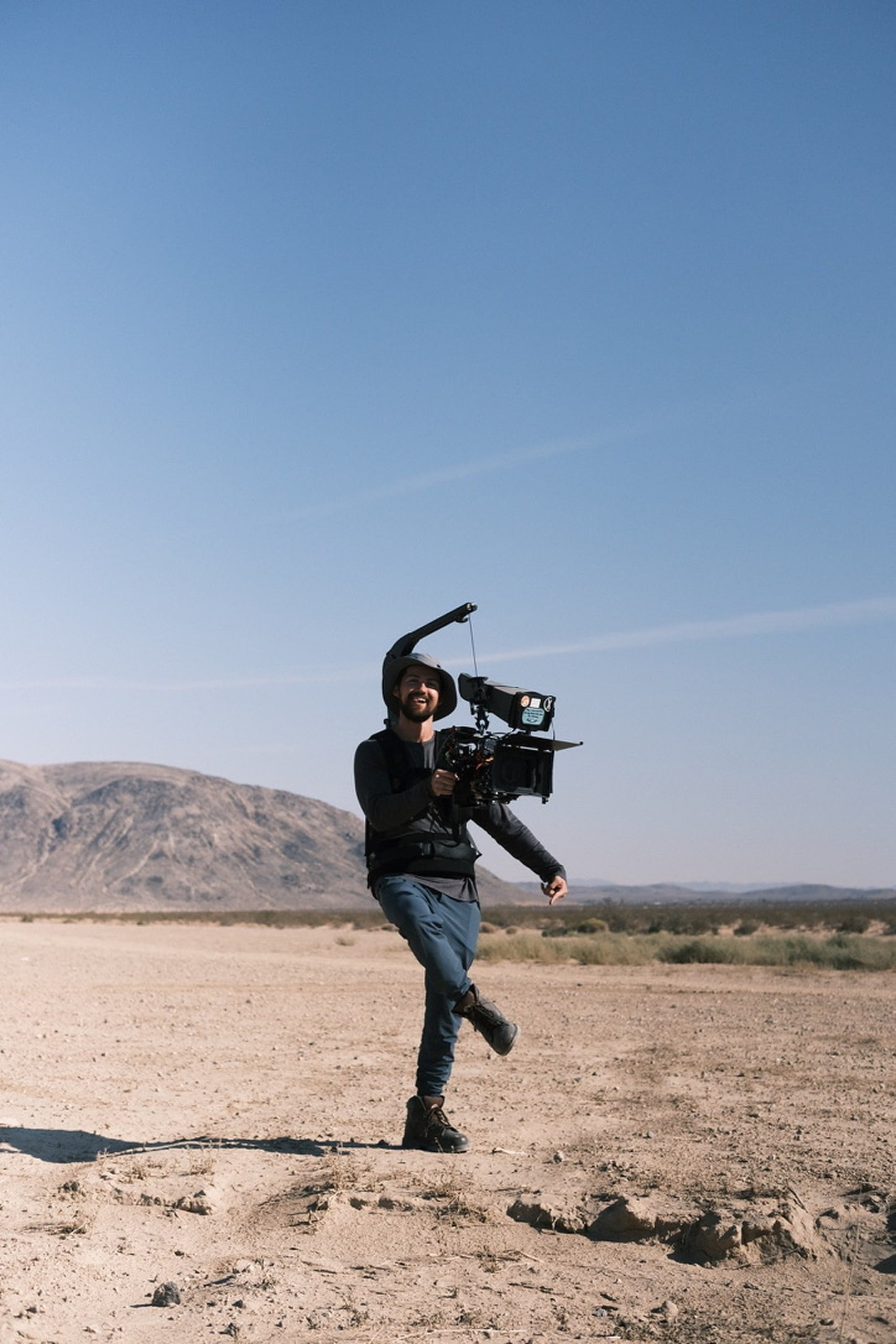 A camera operator mid-stride in the desert, rig on his shoulder, grinning
