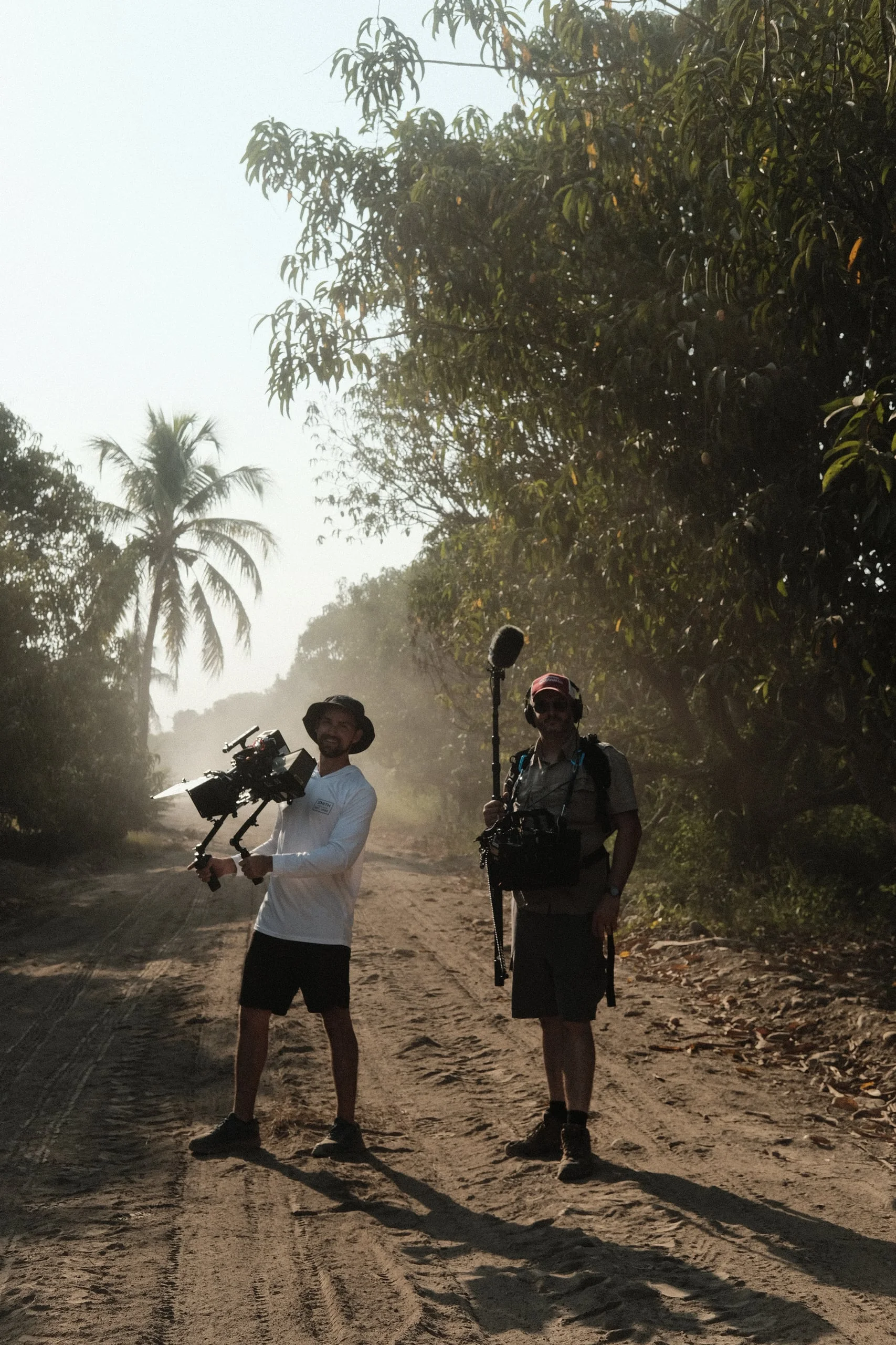A camera operator and sound recordist stand on a dusty road under the trees