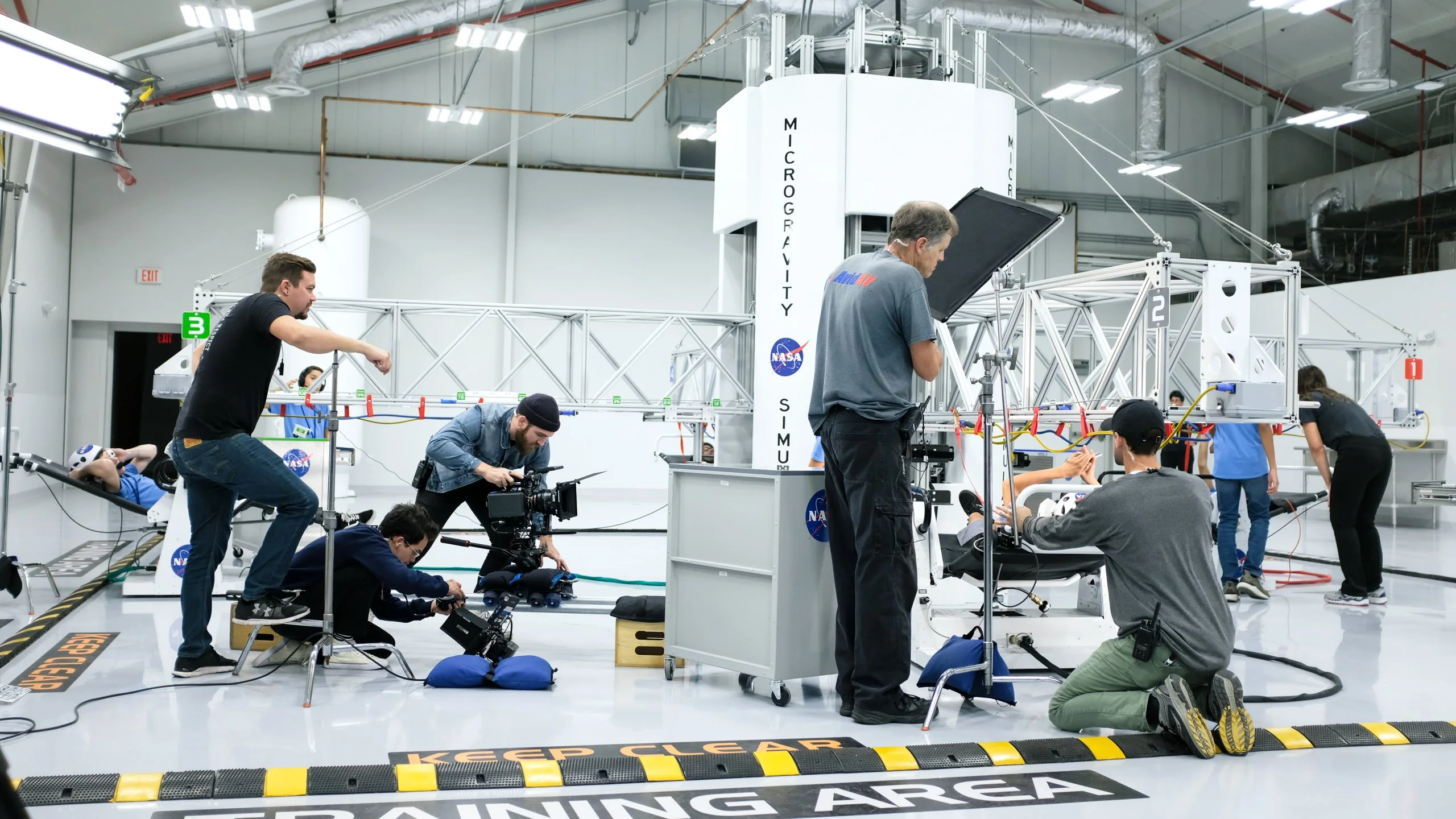 A Spry crew filming on a white soundstage floor marked Training Area, cameras and lights around a tall simulator tower