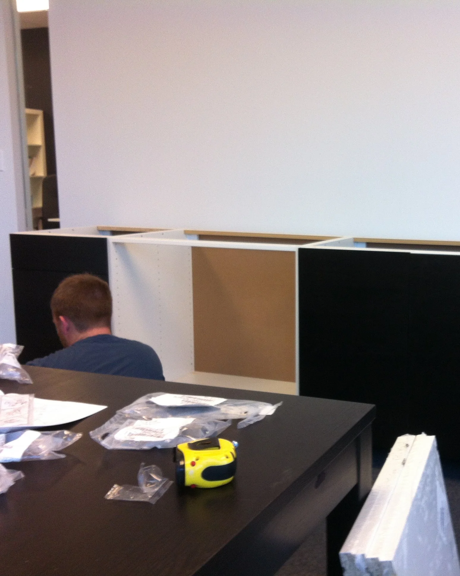 Black cabinets half assembled along a white wall, hardware bags and a tape measure on the table