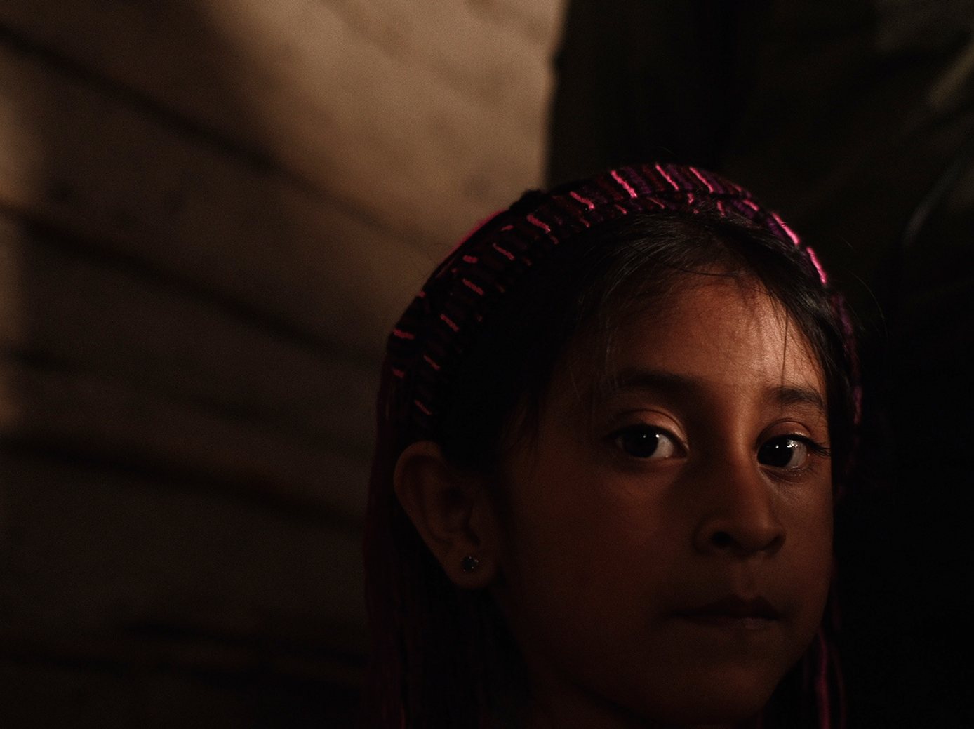 A girl in a woven headband looks into the camera in low light