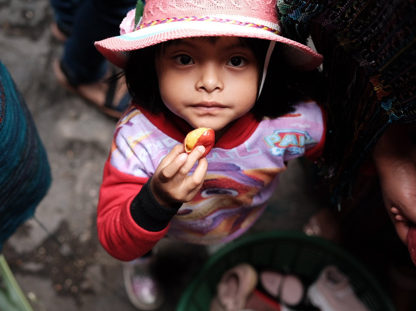 A young girl in a pink sun hat holds up a piece of fruit in a market