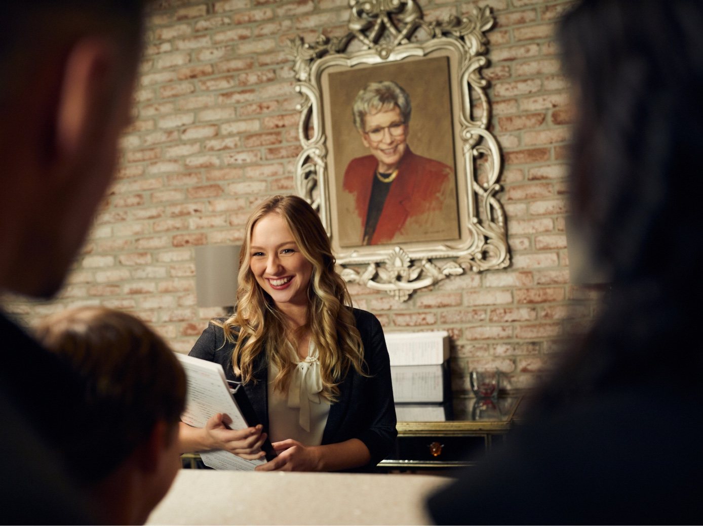 A host smiles at arriving guests beneath a framed portrait of Ruth Fertel