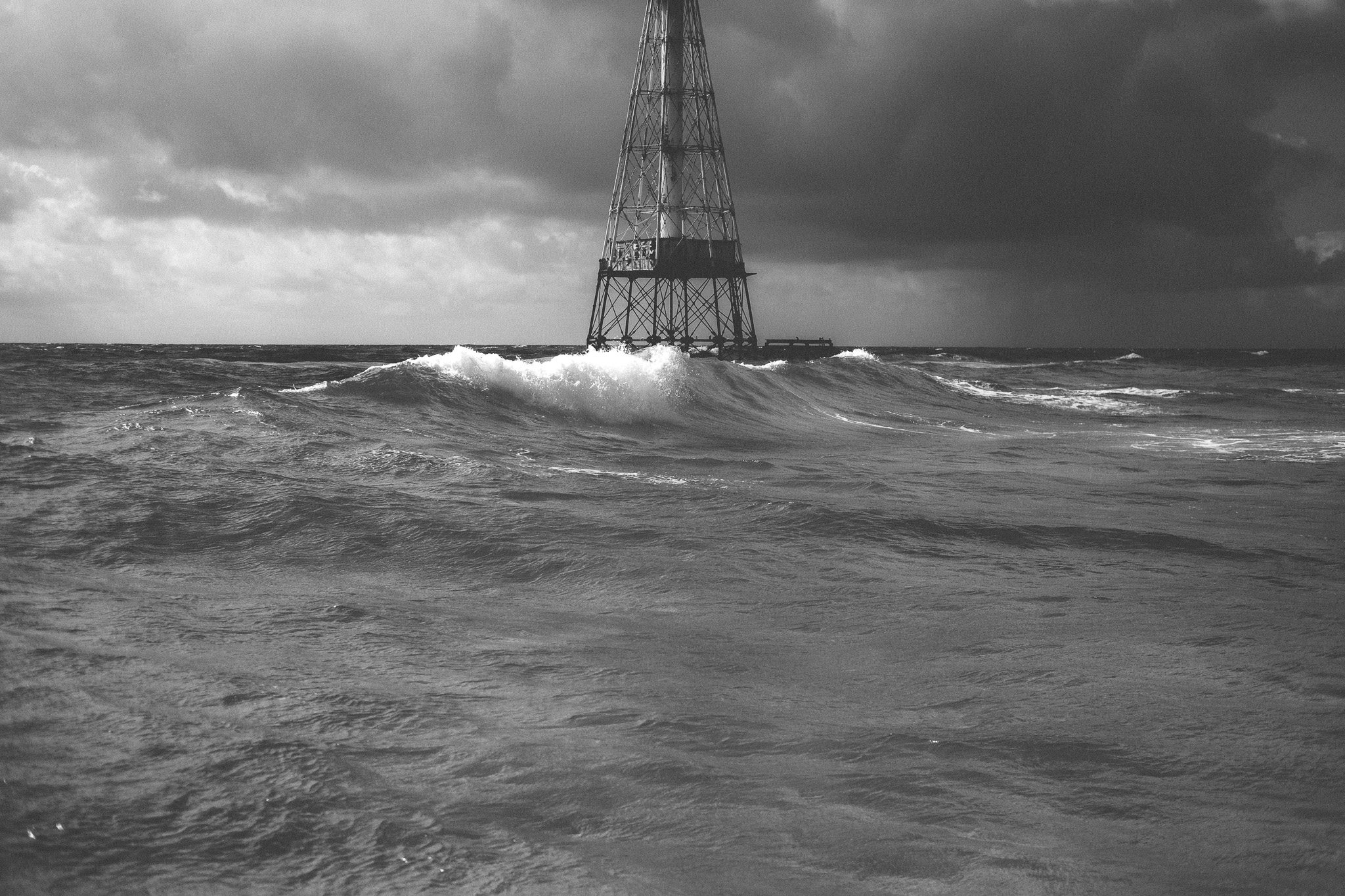 A wave breaks in front of an offshore lighthouse tower under dark storm clouds
