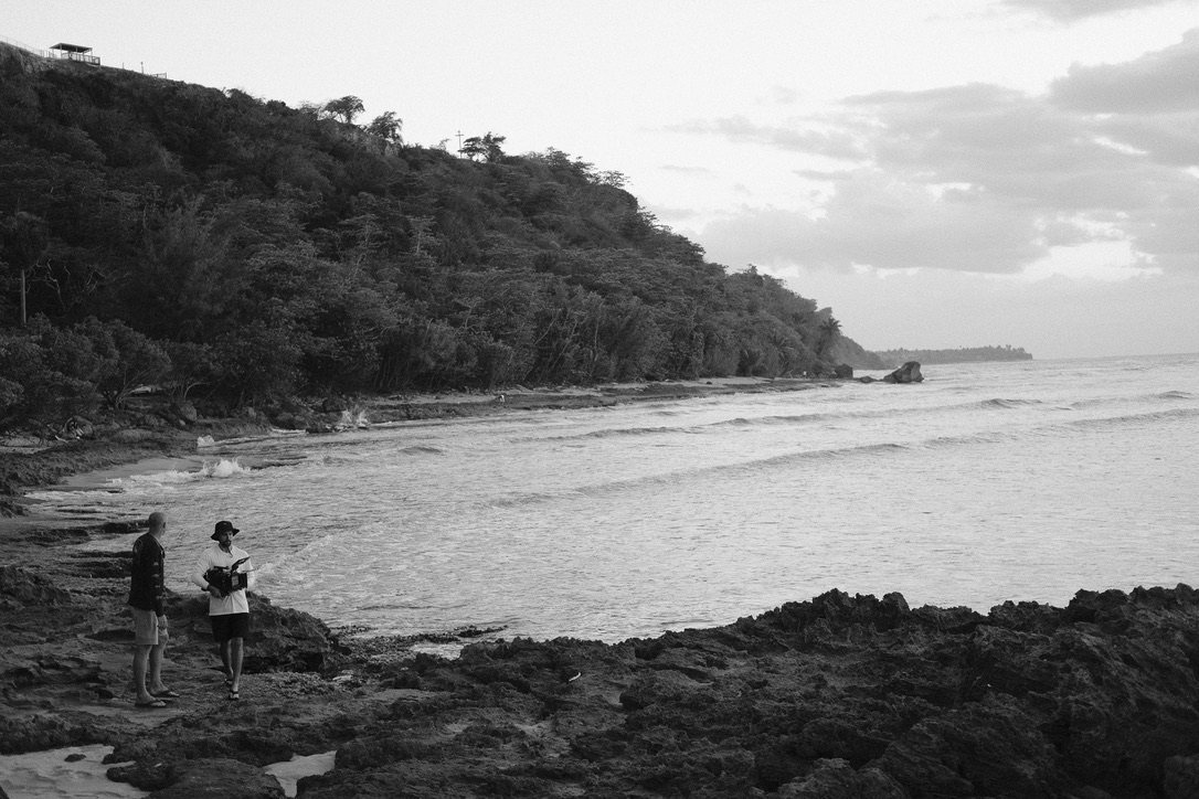 Two crew members on rocks at the water's edge below a wooded hillside