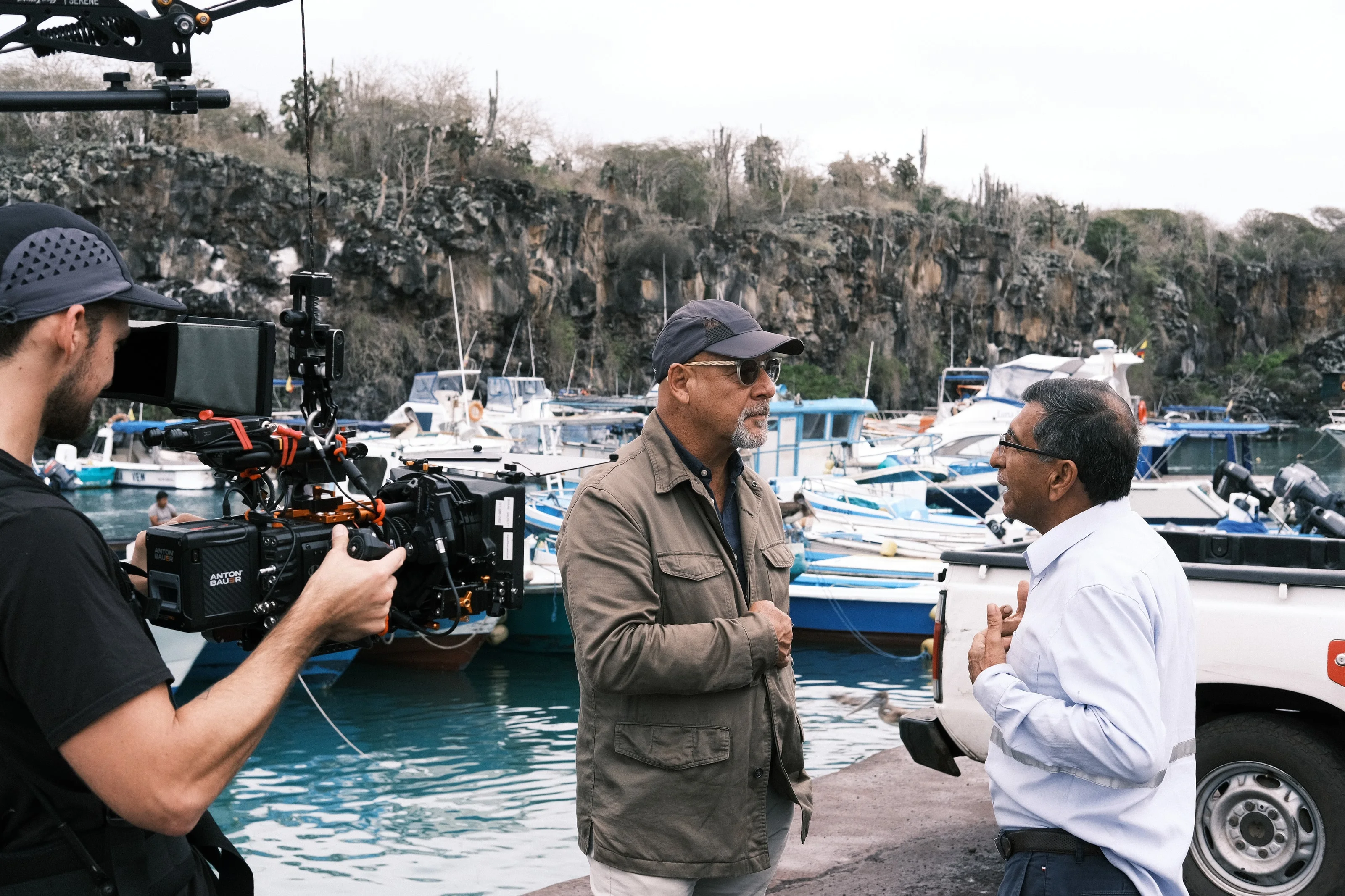 A camera operator films two men talking on a harbor wall lined with small boats
