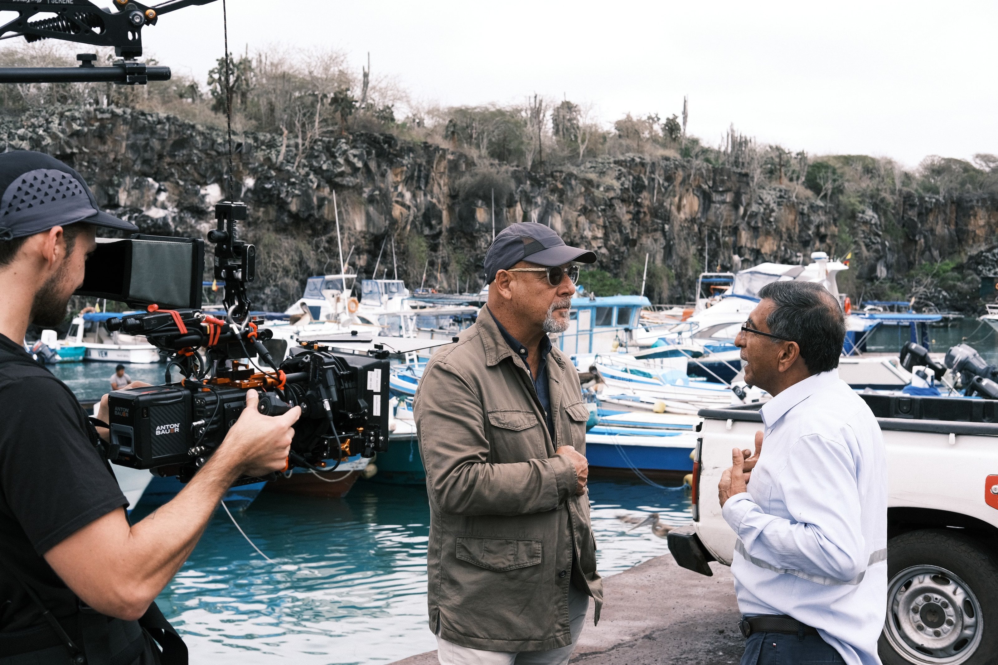 A camera operator films two men talking on a harbor wall lined with small boats