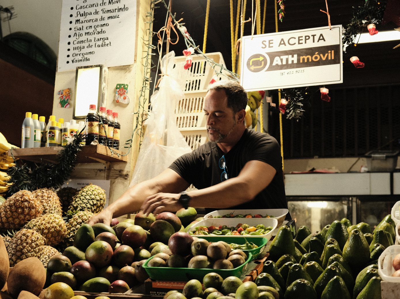 A vendor arranges mangos at a market stall piled with pineapples and avocados