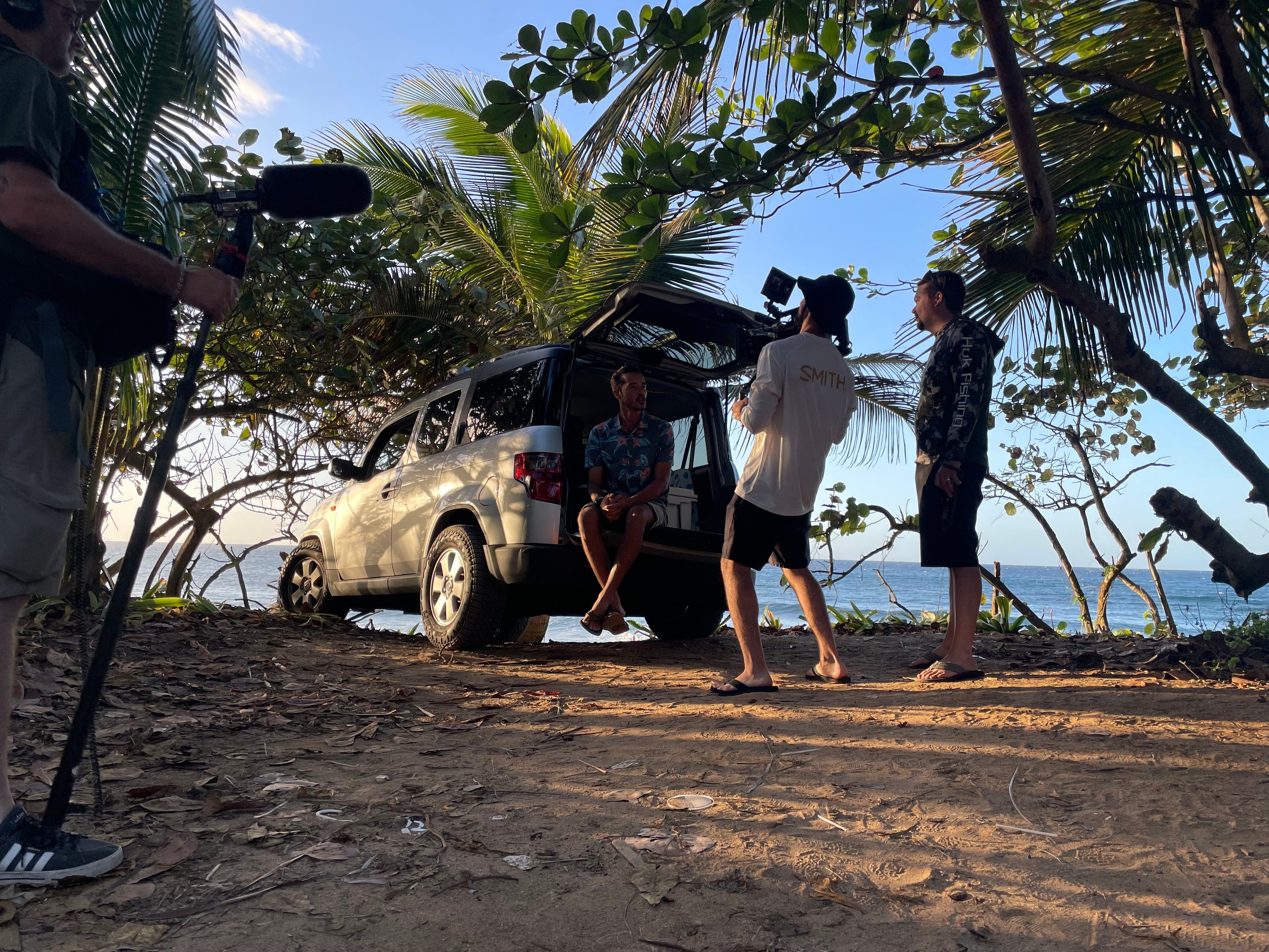 The crew filming an interview beside an SUV under the palms, the ocean behind
