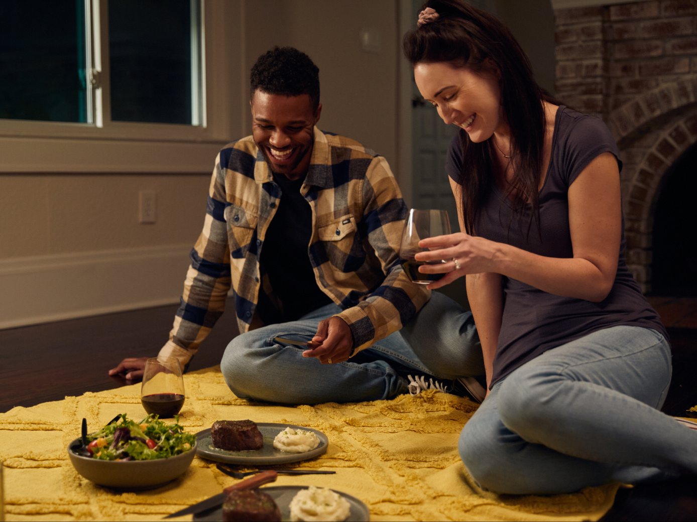 A couple laughs over wine and steak dinners on a yellow blanket spread on a living room floor