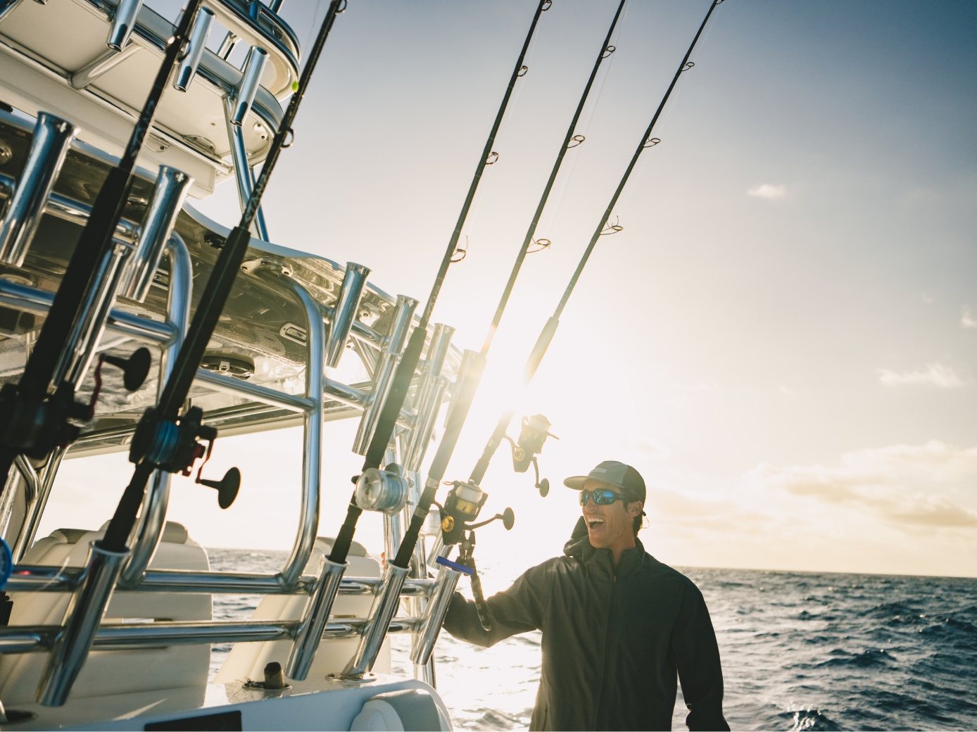 An angler stands among rods in their holders, backlit by the sun offshore