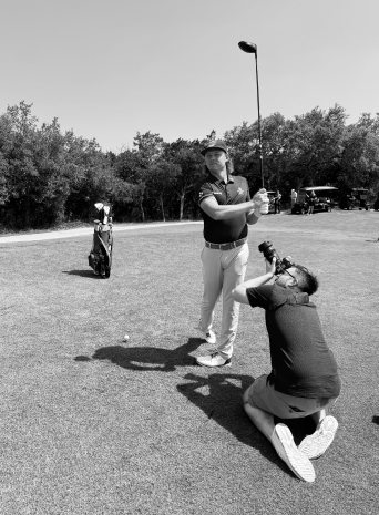 A photographer kneels on the grass shooting up at a golfer mid-swing