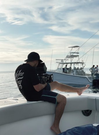 A camera operator sits on the edge of a boat filming another fishing boat alongside
