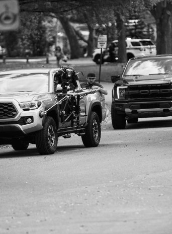 Black and white photo of a camera rig mounted on a pickup truck driving down a tree-lined street