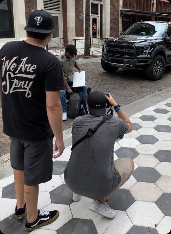A photographer crouches on a tiled sidewalk shooting a man with a backpack as a crew member in a We Are Spry shirt looks on
