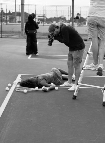 A photographer leans over a player lying on the court among tennis balls while a crew member stands on a ladder