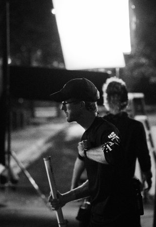 Black and white photo of a crew member in a cap holding a stand at night under a large light