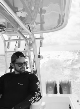 A man in sunglasses and a backwards cap sits at the helm as spray flies behind the boat