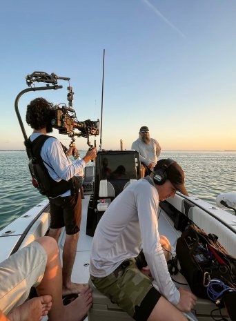 A camera operator with a rigged camera and the sound mixer on a boat at sunset as a bearded captain looks on