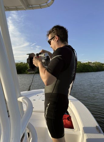 A crew member in a wetsuit and sunglasses readies a camera in a water housing on a boat