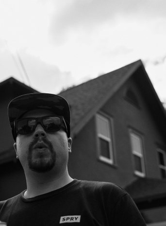 Black and white low-angle portrait of a crew member in sunglasses and a Spry shirt in front of a house