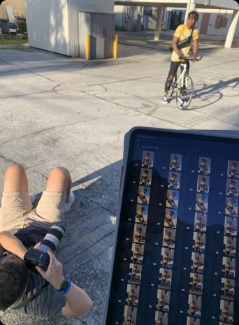 A photographer lies on the pavement shooting a cyclist while a tablet shows a grid of frames
