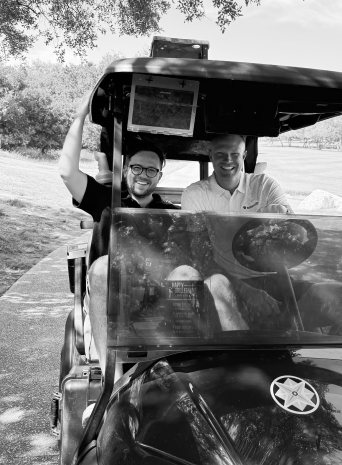 Two men grinning from the front seat of a golf cart