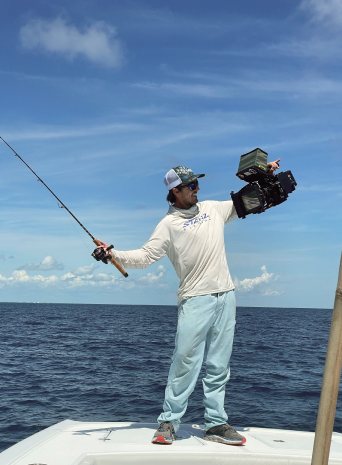 An angler on the bow holds a fishing rod in one hand and a cinema camera in the other