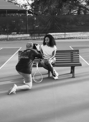 A photographer kneels to shoot a tennis player sitting on a courtside bench with her racquet
