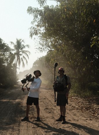 A camera operator and sound recordist on a dusty road under palm trees