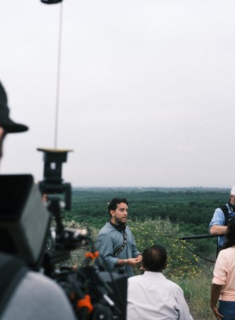 A man talks to the crew on a hill above an orchard while the camera rolls