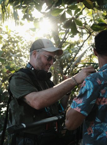 The sound recordist clips a mic onto a man in a floral shirt under the trees