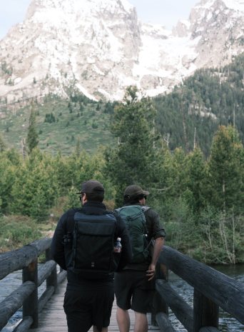 Two crew members with packs crossing a footbridge toward the Tetons