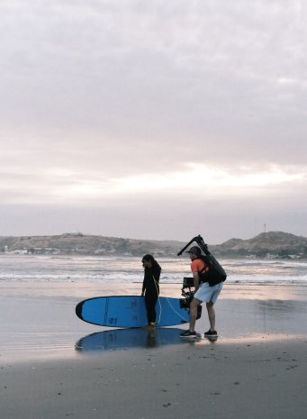A camera operator films a surfer carrying her board on the beach at dusk