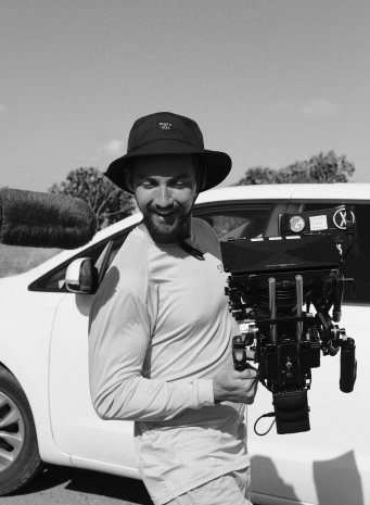 A camera operator in a bucket hat carries the camera past a white car