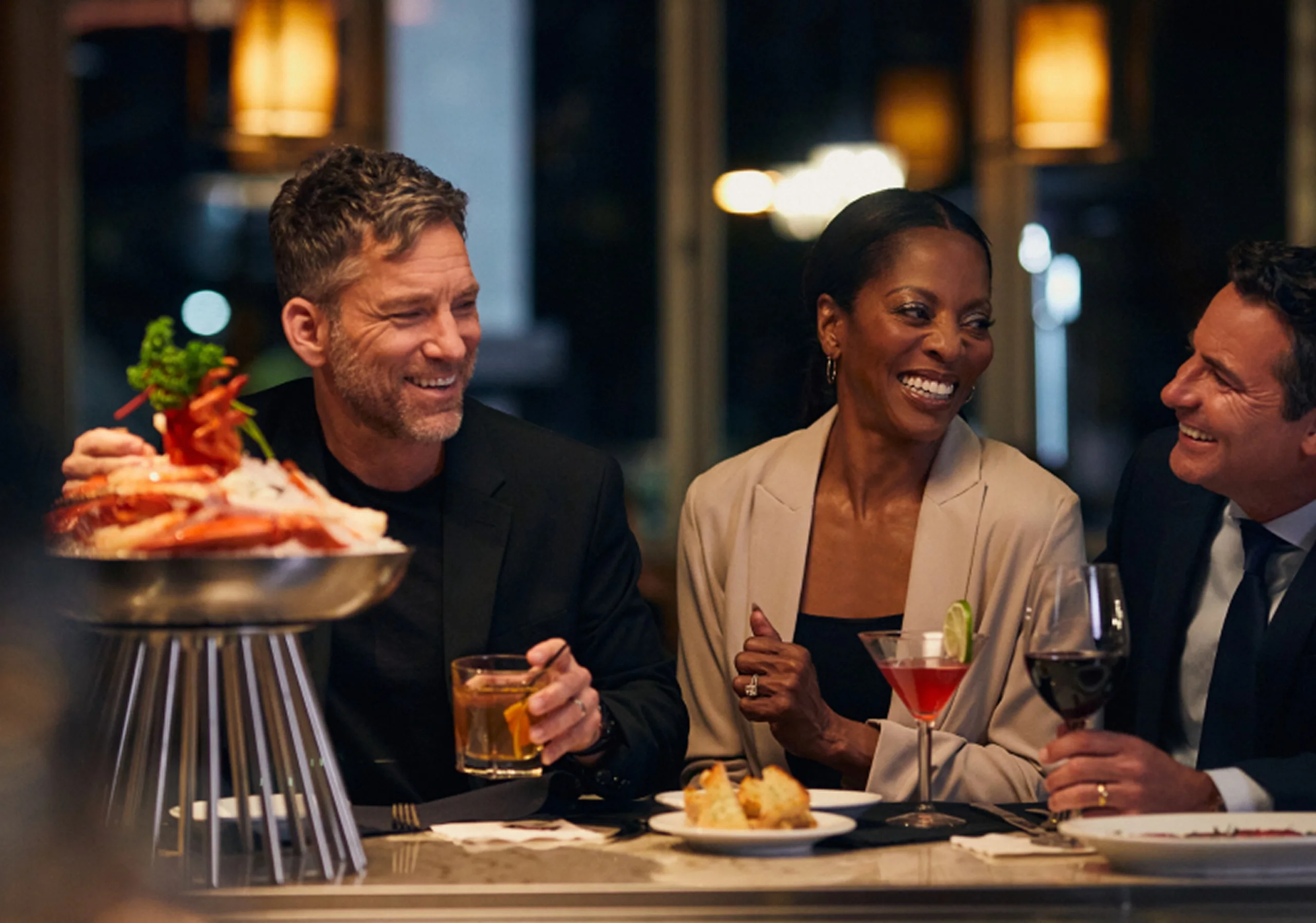 Three diners laugh over cocktails and a seafood tower at Ruth’s Chris