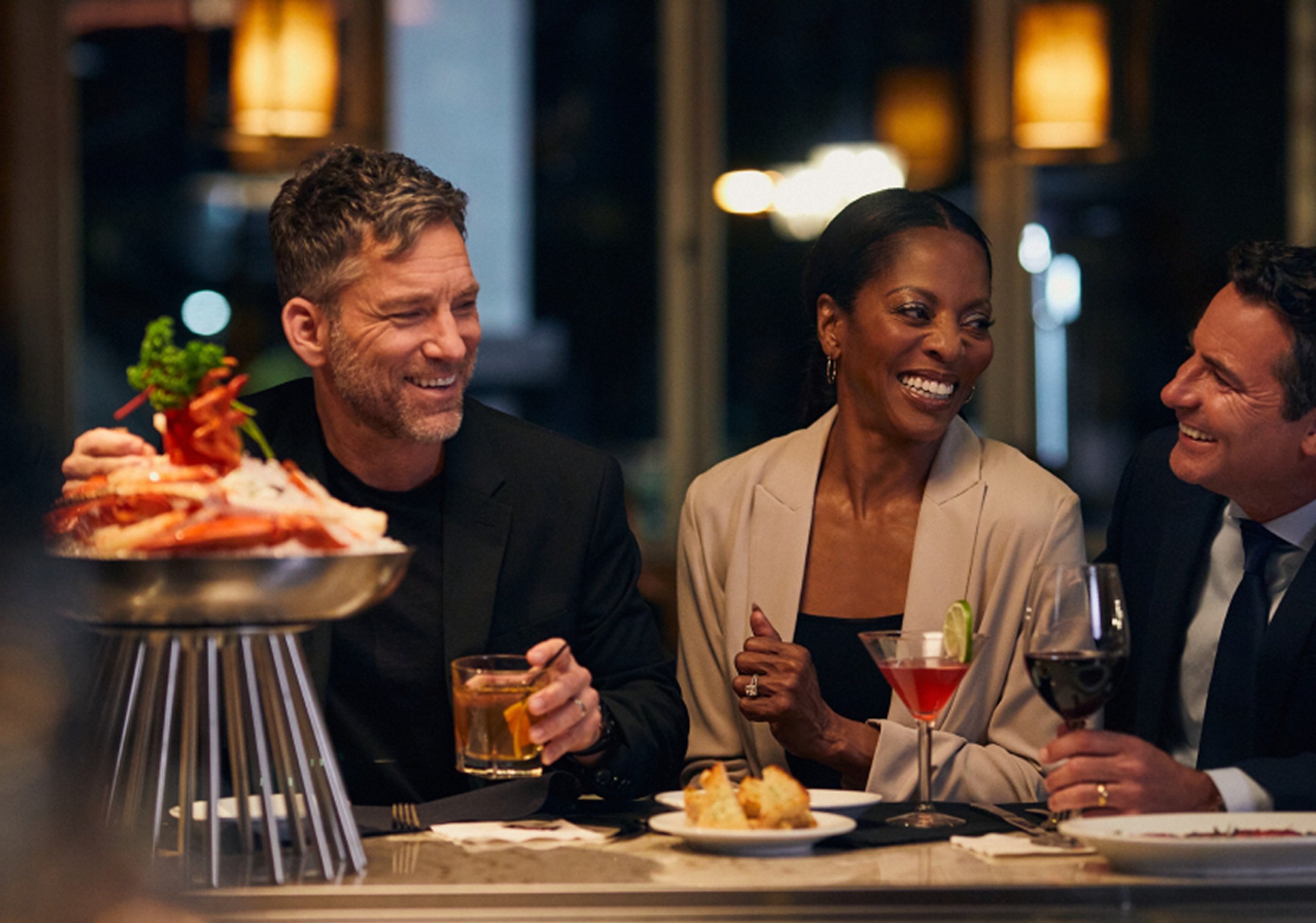 Three friends laugh over drinks and a seafood tower at the bar