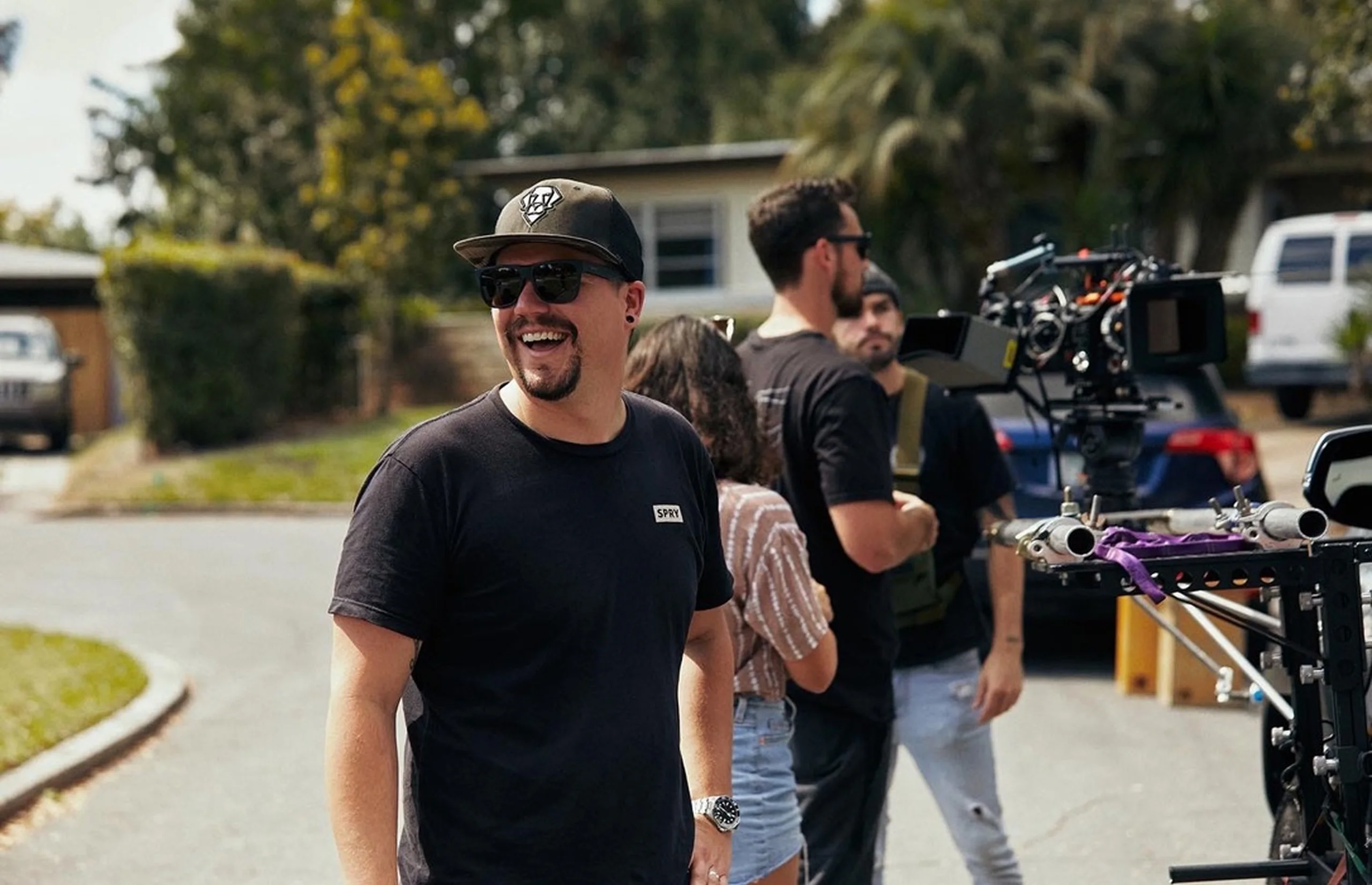 A crew member laughs on a residential street beside a camera on a car rig